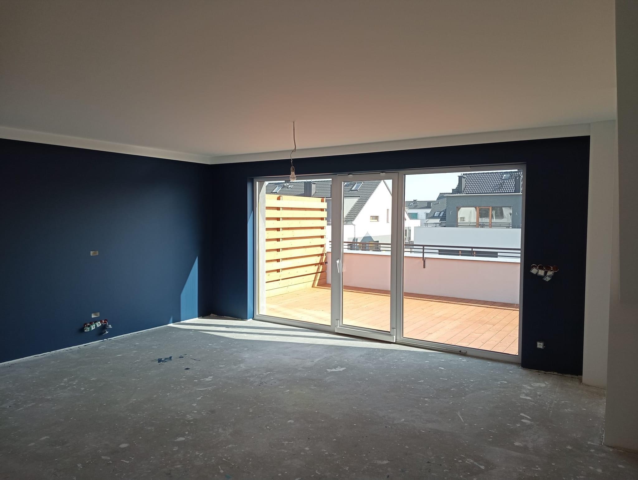 Wnętrze pokoju po malowaniu ścian na ciemny granat, z widocznymi puszkami elektrycznymi i wyjściem na balkon z drewnianą podłogą i balustradą.
