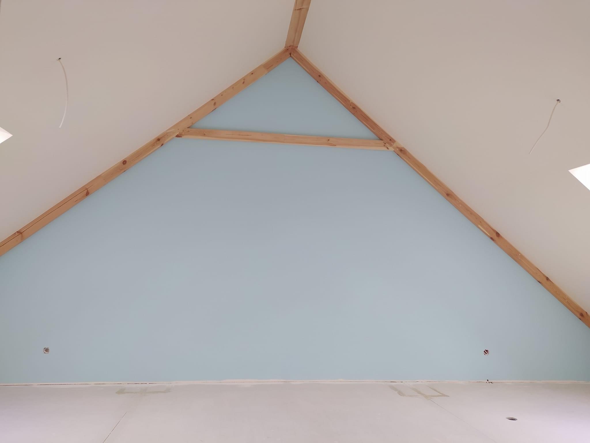 Pomalowana na błękitno ściana szczytowa poddasza z widoczną drewnianą konstrukcją więźby dachowej i wystającymi przewodami elektrycznymi z sufitu.