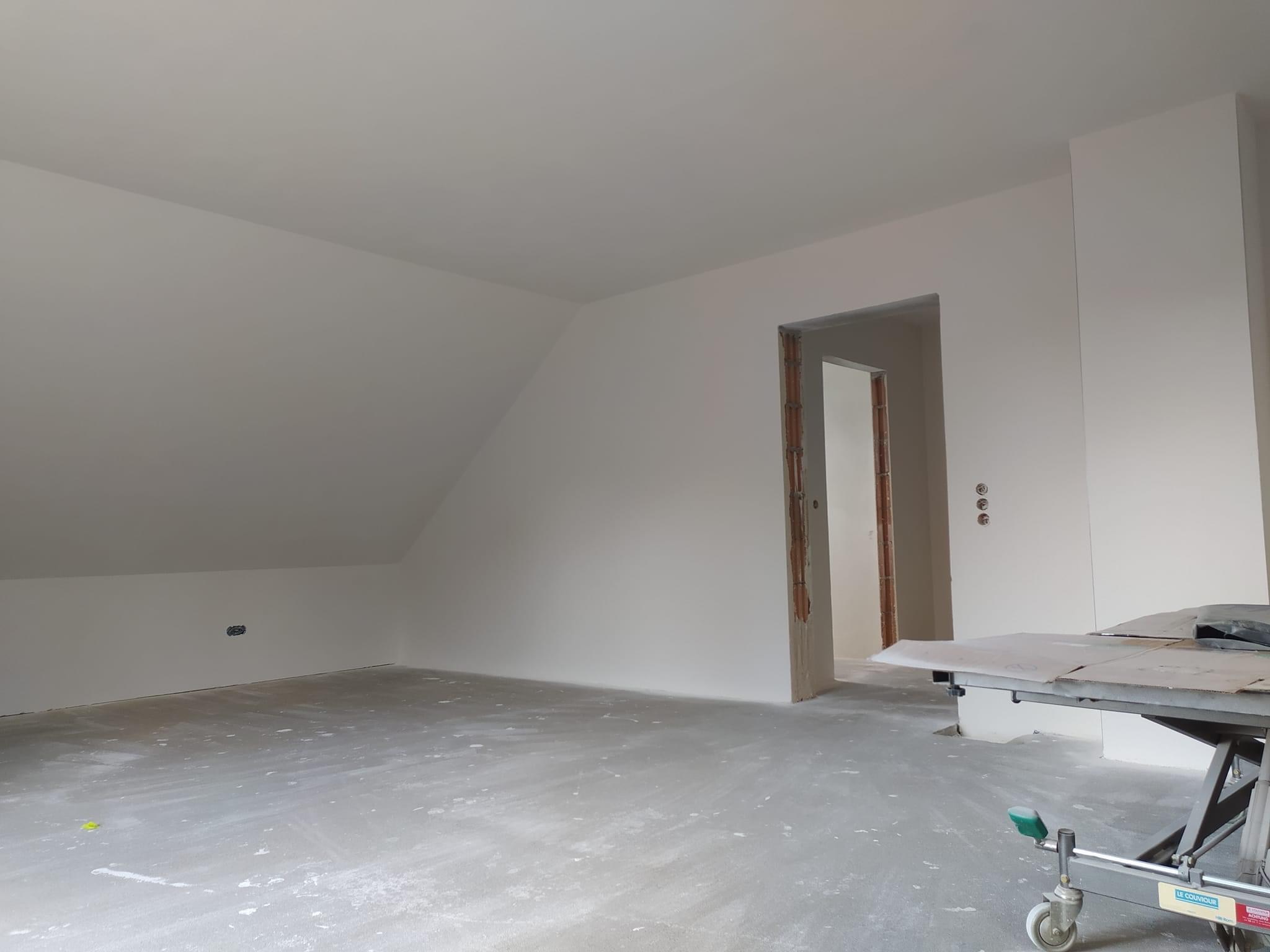 Pokój na poddaszu w trakcie prac wykończeniowych: świeżo położone gładzie na ścianach, widoczny otwór drzwiowy bez ościeżnicy, podłoga pokryta pyłem, w tle widoczny stół roboczy z narzędziami.