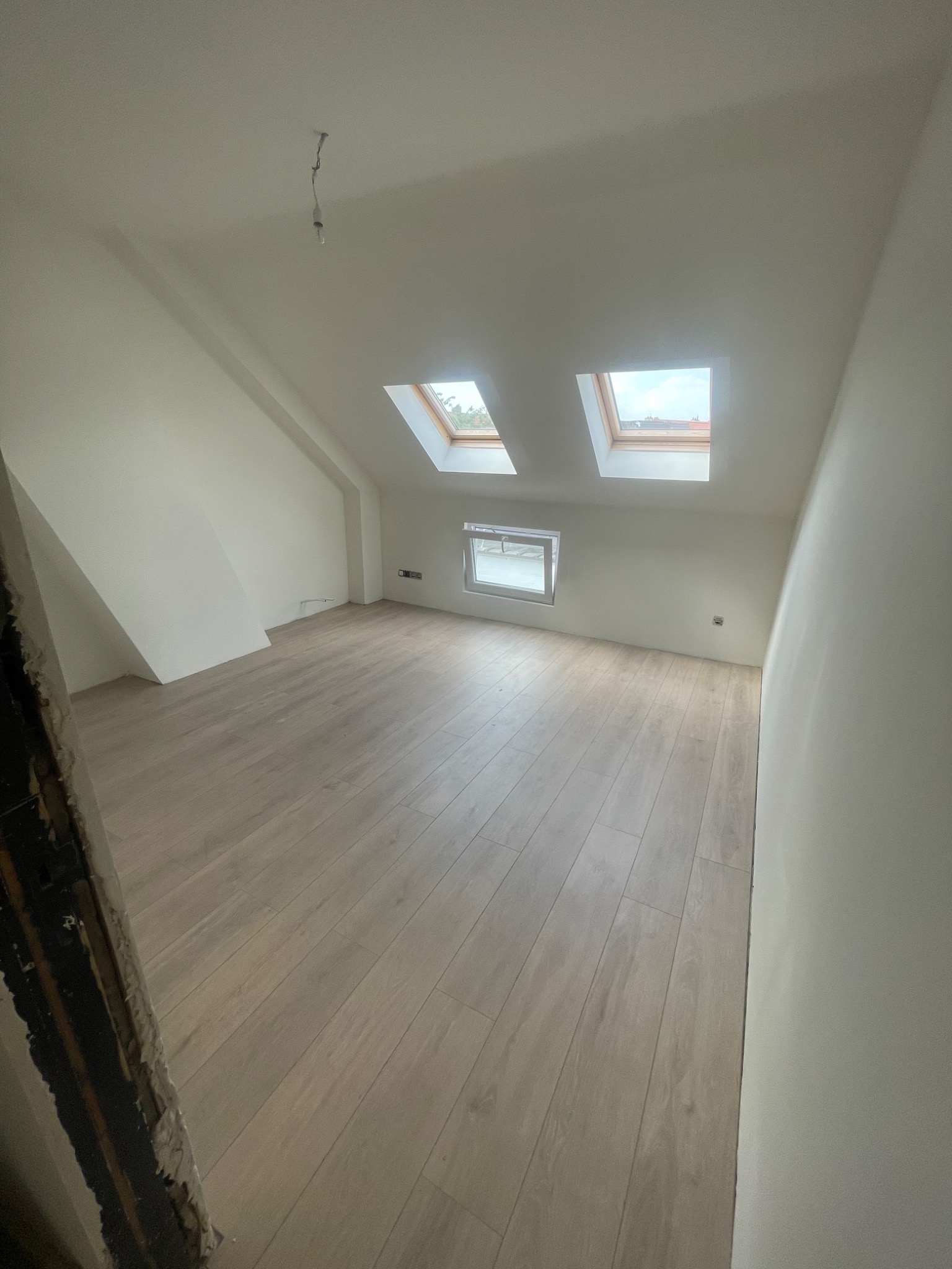 Poddasze po remoncie z jasną podłogą panelową i białymi ścianami, widoczne dwa okna dachowe i jedno okno pionowe, surowy fragment ościeżnicy po lewej stronie.