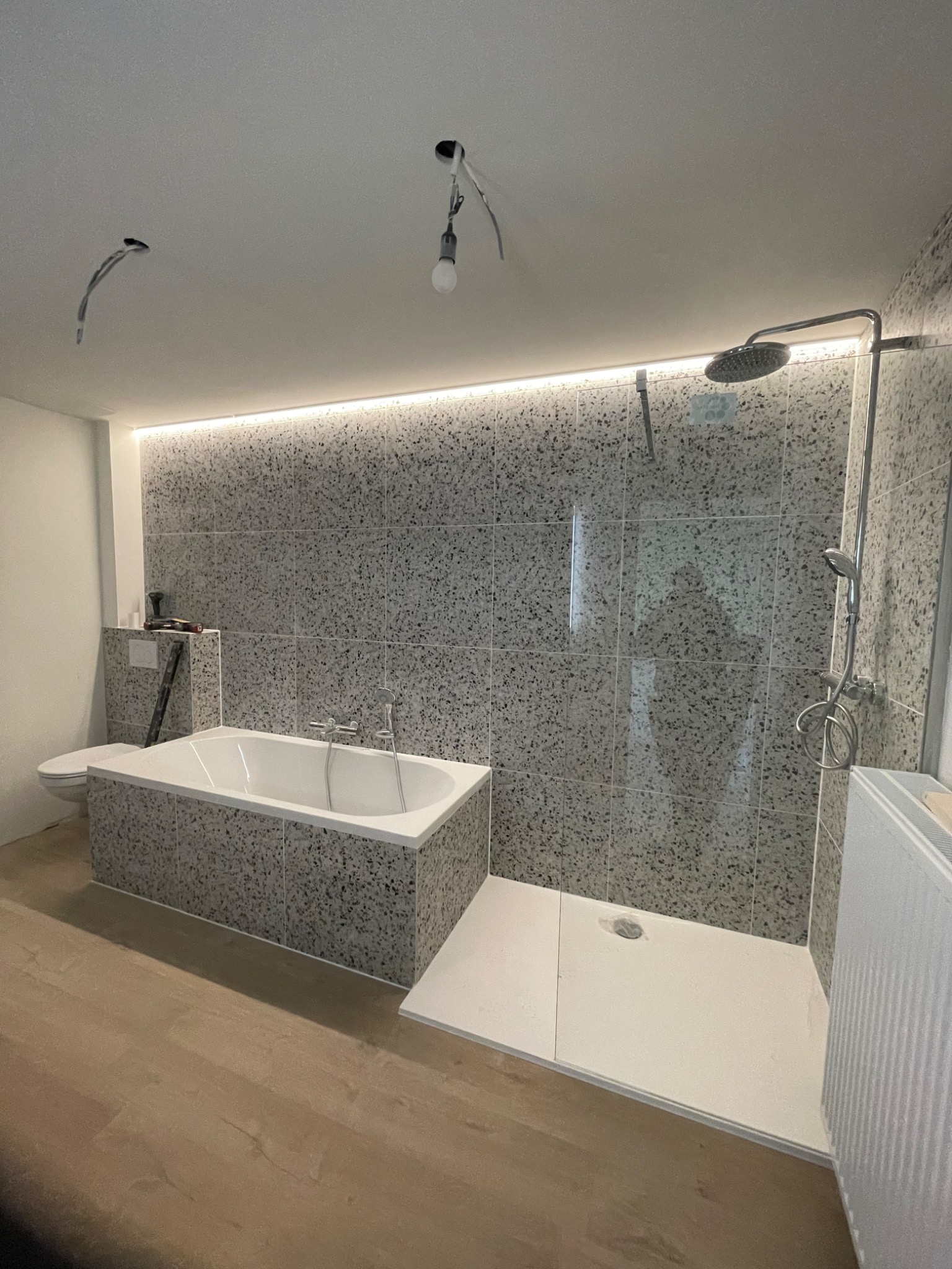 Łazienka w trakcie wykańczania: wanna obudowana płytkami imitującymi lastryko, kabina prysznicowa walk-in z odpływem liniowym, podświetlenie LED nad płytkami, widoczne niedokończone instalacje...