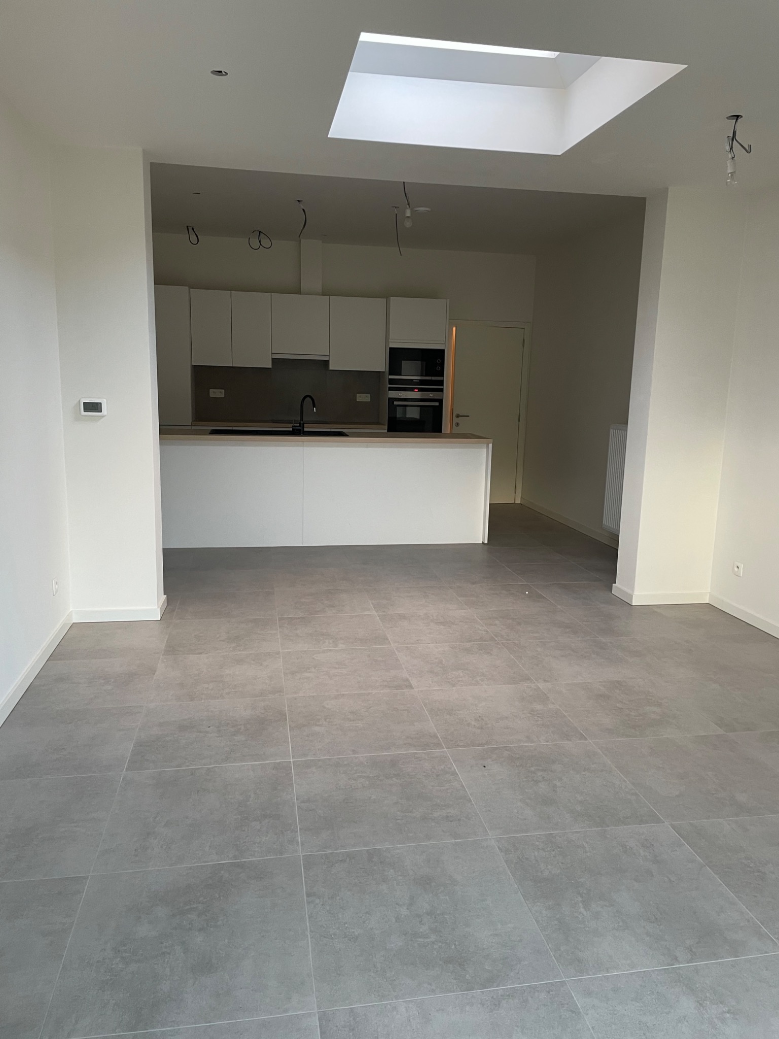 Nowoczesne wnętrze z szarymi płytkami podłogowymi, białą kuchnią w tle i świetlikiem sufitowym, widoczne niedokończone instalacje elektryczne.