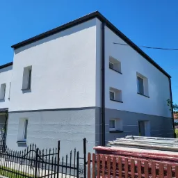 Dwukondygnacyjny dom z elewacją w kolorze szaro-białym, z widocznym ogrodzeniem i stertą materiałów budowlanych na pierwszym planie.