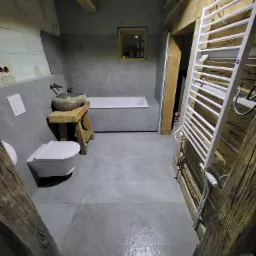 Surowa łazienka z szarymi płytkami, drewnianymi belkami stropowymi i umywalką przypominającą kamień na drewnianym postumencie, grzejnik drabinkowy na drewnianej ścianie.