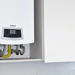 Biały piec gazowy Termet z wyświetlaczem w szafce kuchennej, widoczne podłączenia rur gazowych i wodnych.