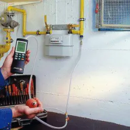 Sprawdzanie szczelności instalacji gazowej za pomocą elektronicznego miernika i pompki ciśnieniowej, widoczne żółte rury i gazomierz na białej ścianie, skrzynka z narzędziami obok.