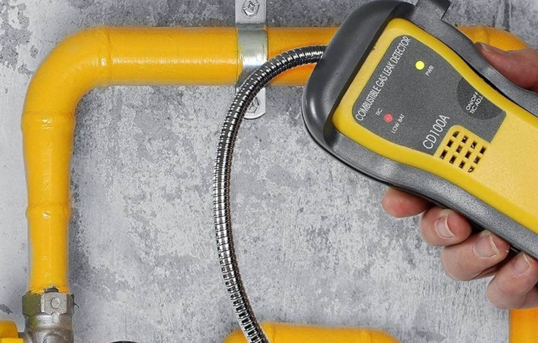 Sprawdzanie szczelności żółtej instalacji gazowej za pomocą detektora nieszczelności gazu, model CD100A, na tle szarej ściany.