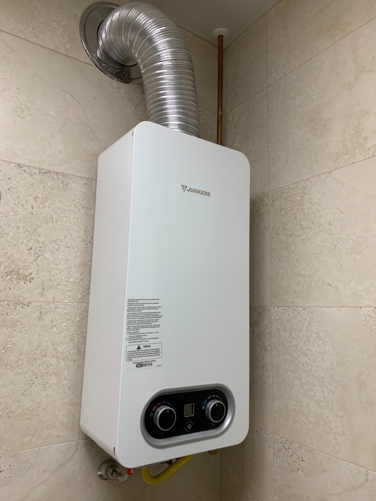 Biały piec gazowy Junkers zamontowany na beżowej ścianie z płytek, widoczny przewód odprowadzający spaliny i rurka doprowadzająca wodę.