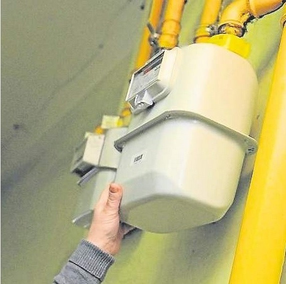 Ręka przytrzymuje obudowę gazomierza zamontowanego na ścianie, widoczne żółte rury instalacji gazowej.