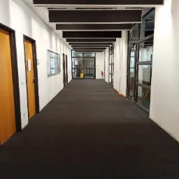 Długi korytarz biurowy z ciemną wykładziną dywanową, białymi ścianami, rzędem drzwi po lewej stronie i przeszklonymi ścianami po prawej, zakończony przeszklonymi drzwiami.