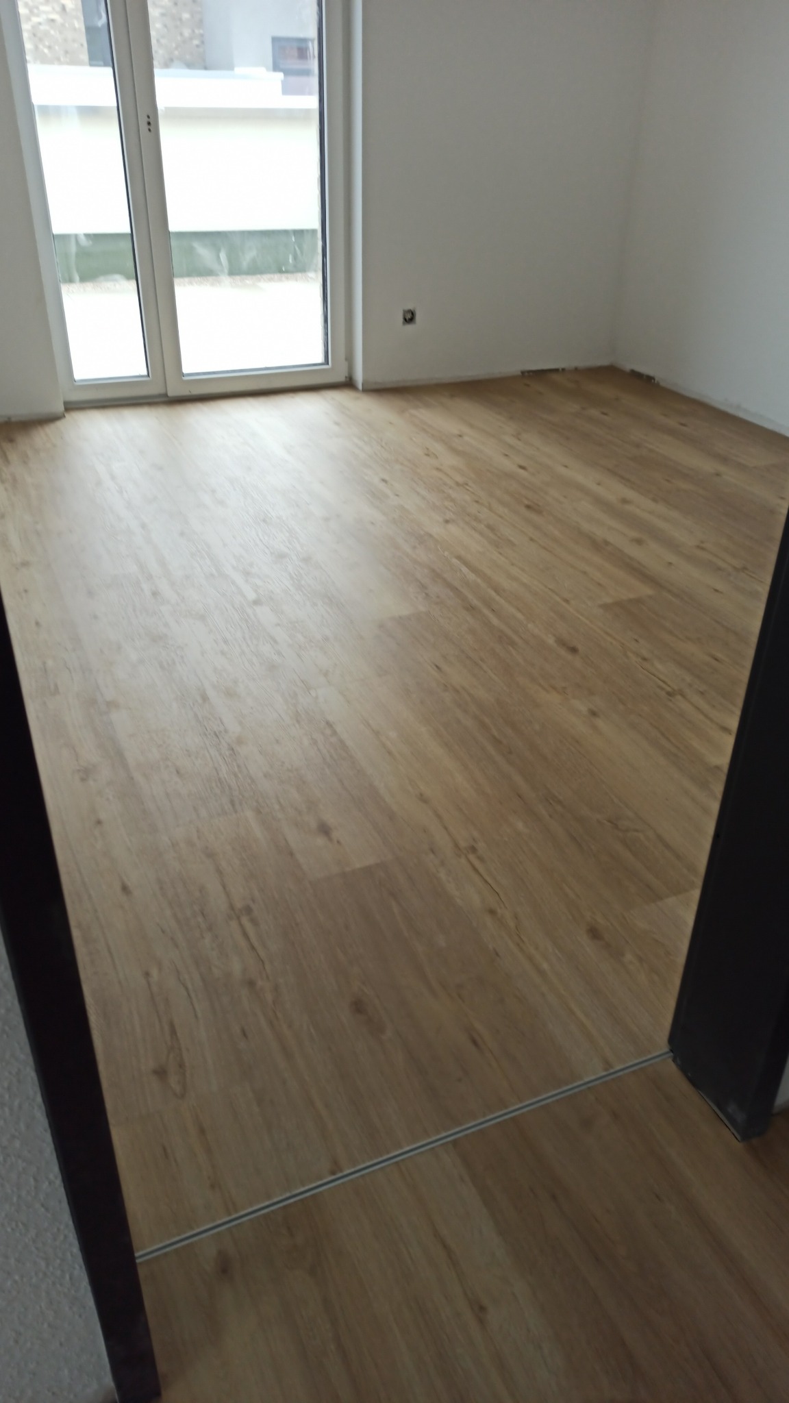 Ułożona podłoga z paneli w jasnym odcieniu drewna, widoczna listwa progowa przy przejściu do innego pomieszczenia, ściany w kolorze białym, widoczne okno balkonowe.