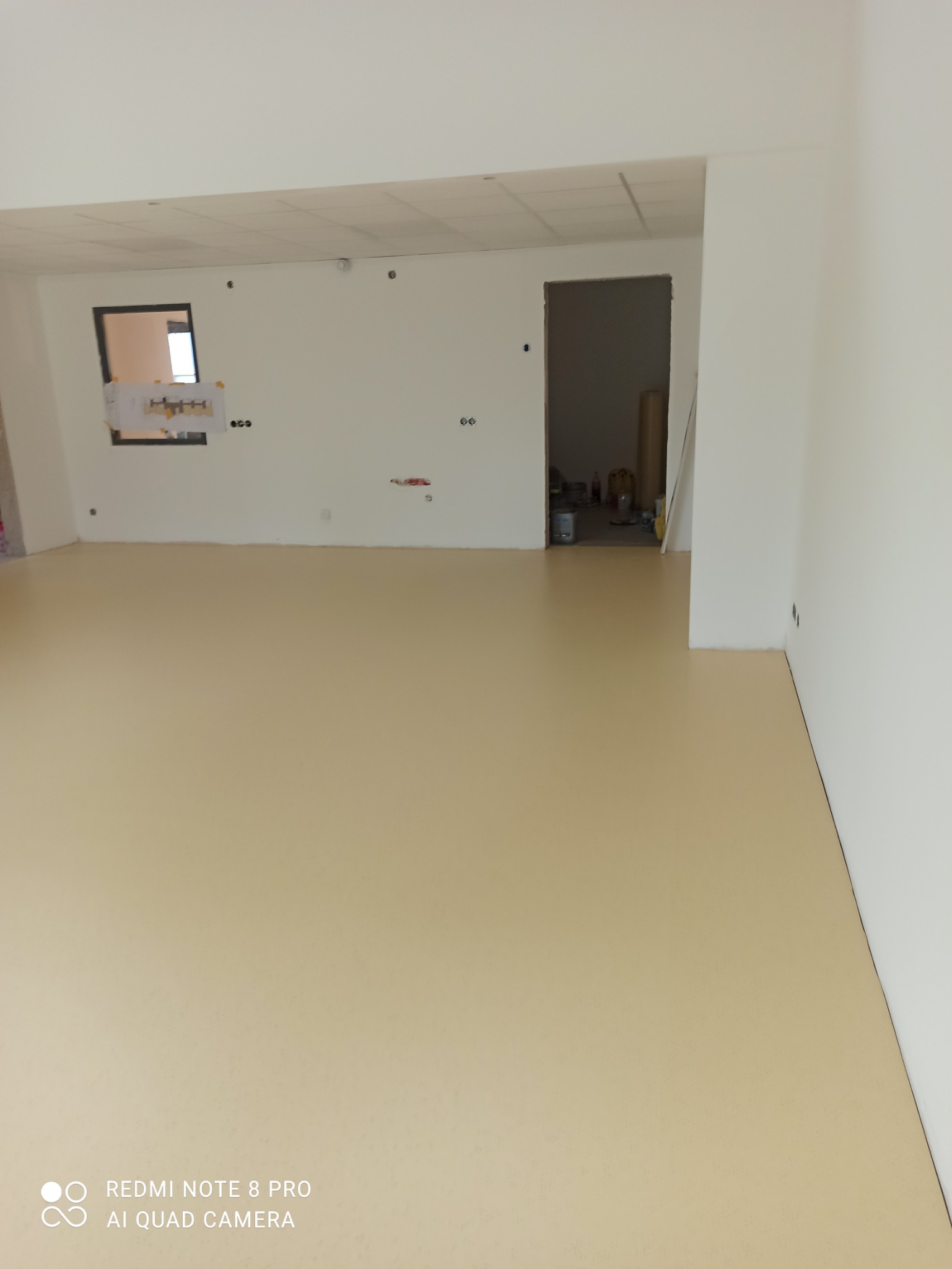 Jasna, beżowa podłoga w surowym pomieszczeniu z białymi ścianami, widoczne puszki i rolki w przejściu do sąsiedniego pokoju, instalacja elektryczna na ścianie.