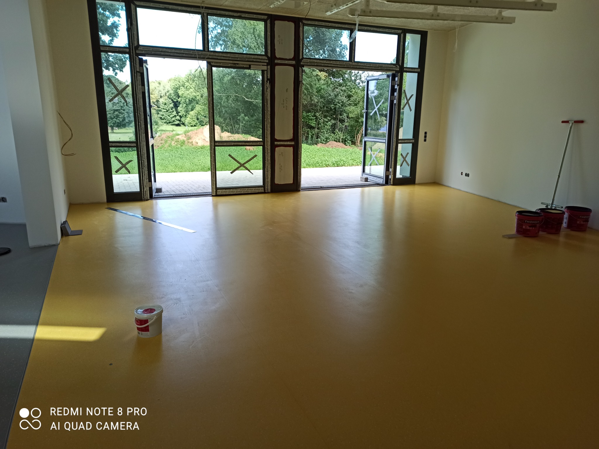 Żółta, gładka wylewka samopoziomująca w jasnym pomieszczeniu z dużymi oknami i widokiem na zieleń, obok ściany stoją puszki z preparatem i narzędzie do rozprowadzania.