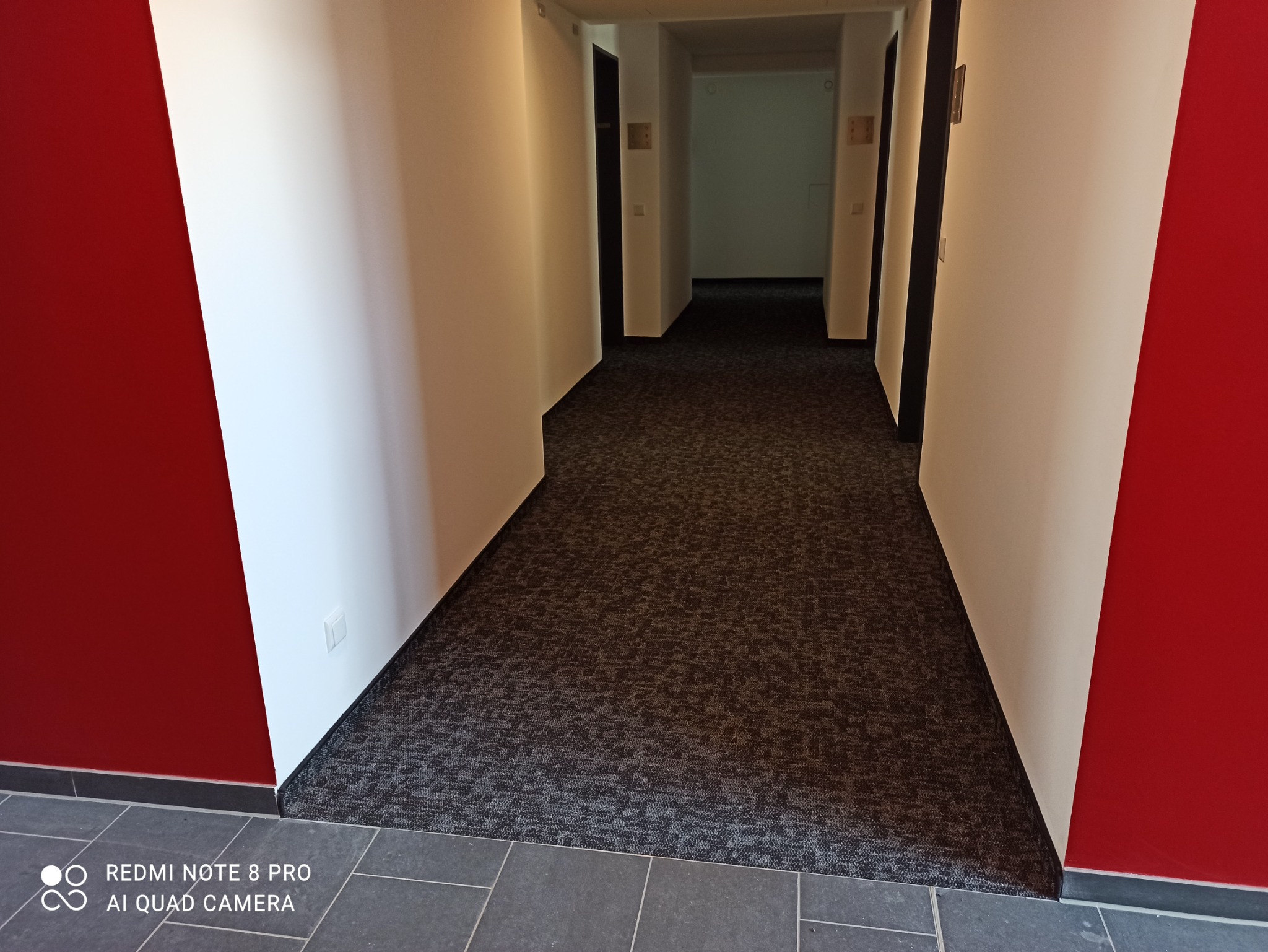 Korytarz hotelowy z ciemną wykładziną dywanową o drobnym wzorze, czerwonymi i białymi ścianami oraz szarą podłogą u dołu kadru.