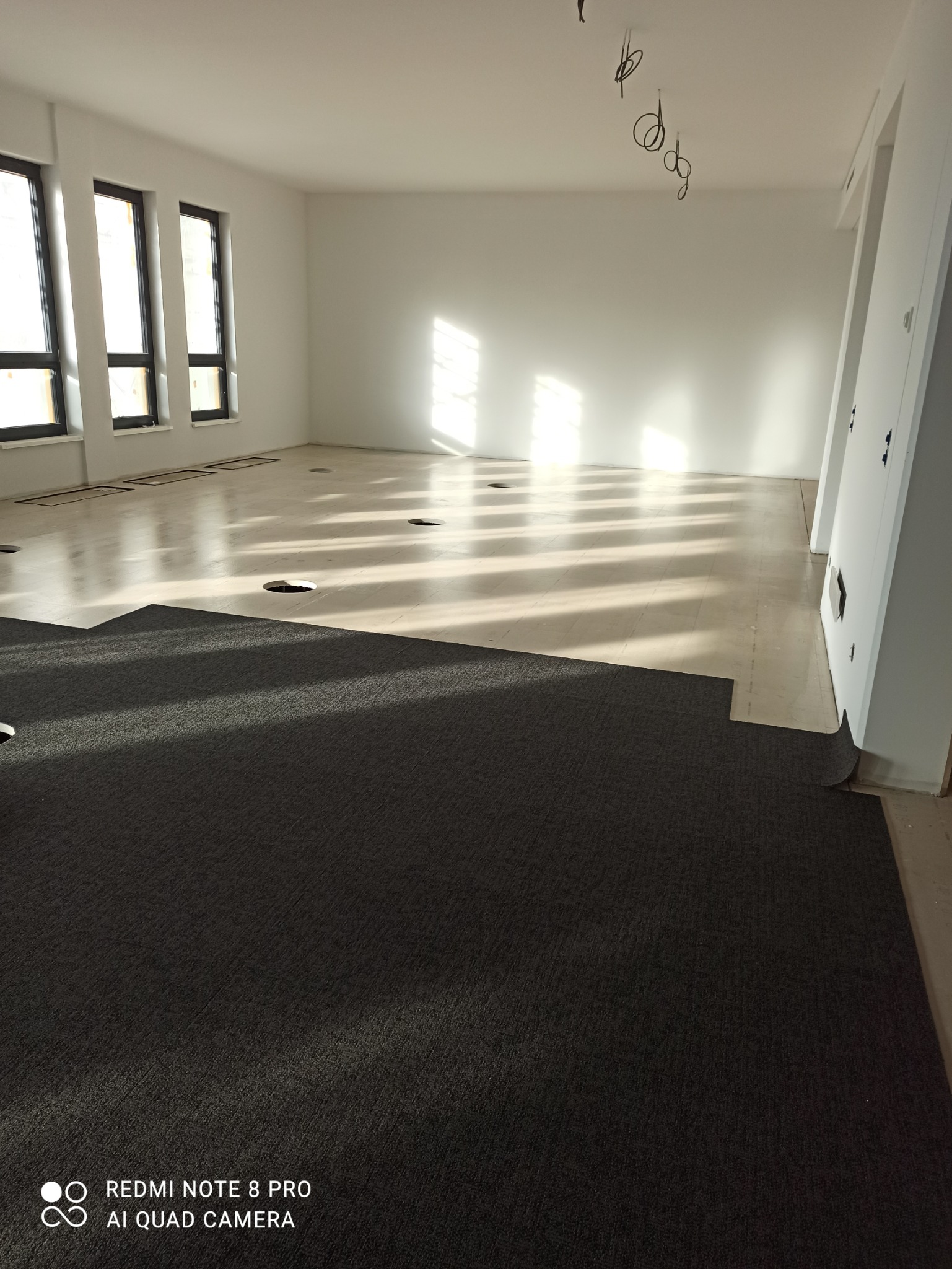 Wnętrze w trakcie montażu wykładziny dywanowej w dużej sali z oknami, widoczne fragmenty podłogi z otworami i niedokończone krawędzie.