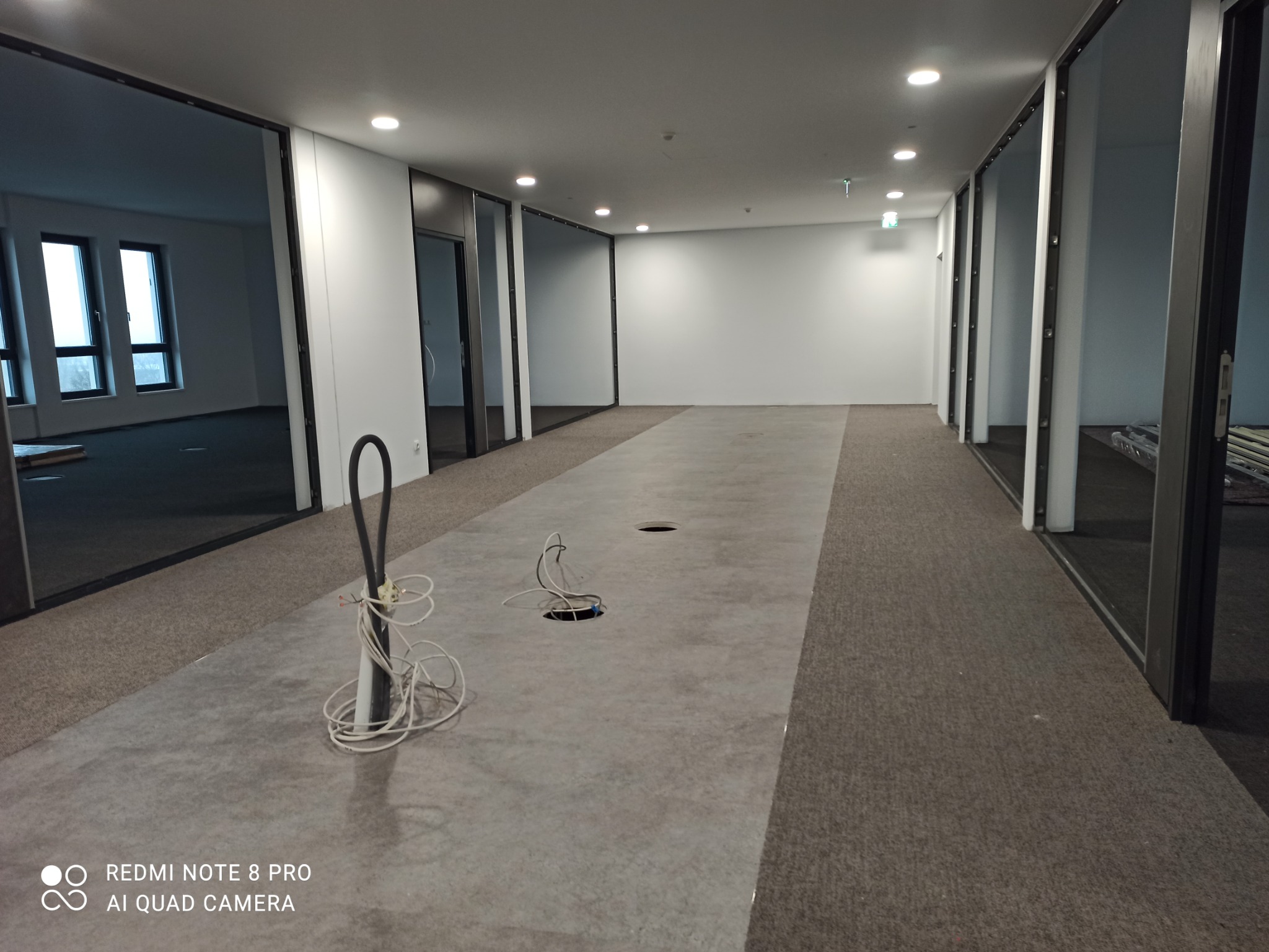 Przygotowanie podłoża pod nową podłogę w biurze: widoczne otwory w wylewce pod instalacje, częściowo położona wykładzina dywanowa, widoczne kable i rury instalacyjne, przeszklone ściany biurowe.