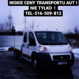 Biała laweta Fiat Ducato z tablicą rejestracyjną LU 402MT, zaparkowana na mokrym, ciemnym asfalcie w pochmurny dzień, z widocznym napisem 'LAWETA 24H Chełm niskie ceny transportu aut i nie tylko!