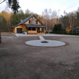 Nowoczesny drewniany dom z szarą stolarką i dachem, otoczony rozległym ogrodem z okrągłym miejscem na ognisko wysypanym żwirem, ścieżką i tujami wzdłuż ogrodzenia.