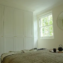 Jasna sypialnia z dużą, wbudowaną szafą z czterema drzwiami, okrągłym lustrem na ścianie i łóżkiem z czajnikiem i filiżankami na tacy.