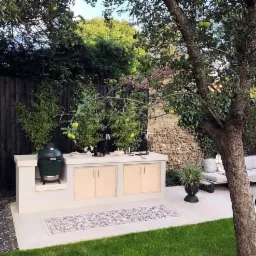 Zewnętrzna kuchnia z blatem z jasnego betonu, szafkami z jasnego drewna, zielonym grillem ceramicznym i dywanem na tarasie, w tle czarny płot i drzewo owocowe.