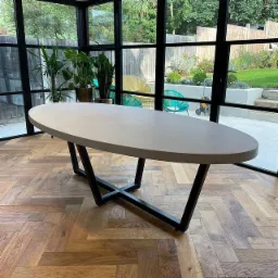 Owalny stół z szarego blatu i czarną, metalową podstawą krzyżową, ustawiony na parkiecie w jodełkę, z widokiem na ogród przez duże okna w czarnych ramach.