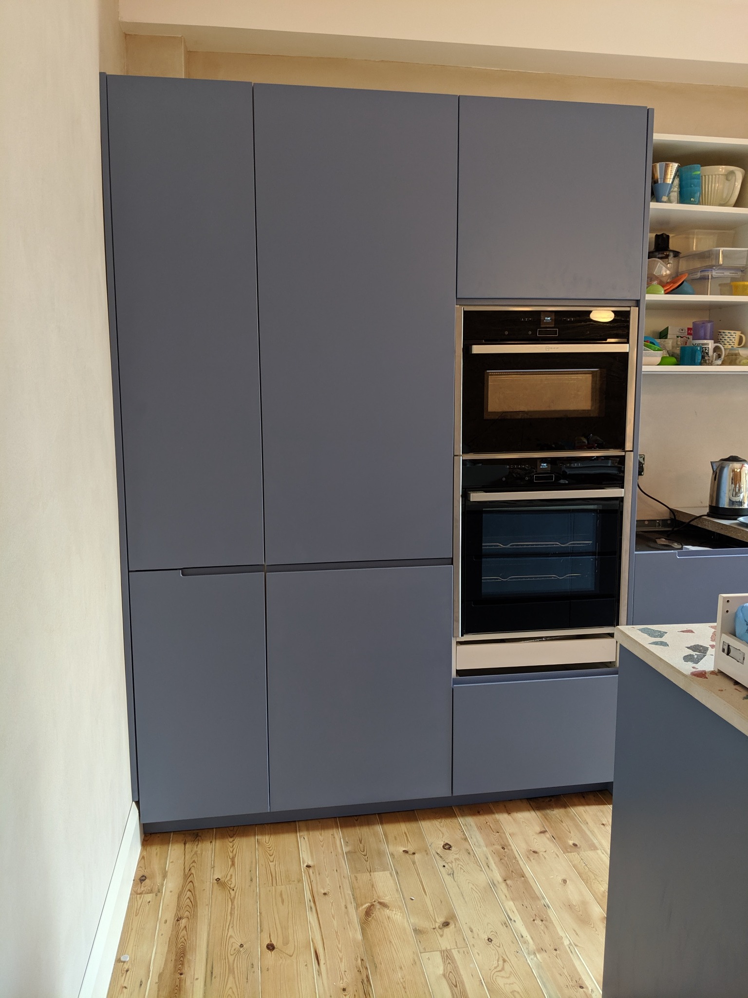 Szara zabudowa kuchenna z piekarnikiem i mikrofalówką wkomponowanymi w szafę, widok na drewnianą podłogę i fragment blatu.