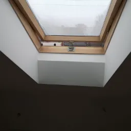 Wnętrze z oknem dachowym z drewnianą ramą, widok na zewnątrz w deszczowy dzień, ściany pomalowane na biało, sufit w odcieniach szarości.
