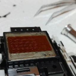 Głowica drukująca z widocznymi dyszami, częściowo zdemontowana, obok narzędzia serwisowe i chusteczka czyszcząca.