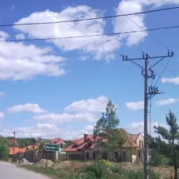 Pejzaż wiejski z widokiem na słupy energetyczne, domy z czerwonymi dachami i znak drogowy z nazwą miejscowości 'Masłów II' pod błękitnym niebem z białymi chmurami.