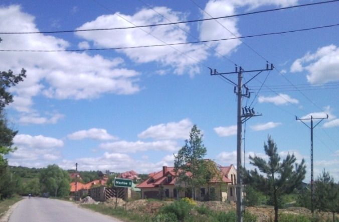 Pejzaż wiejski z widokiem na słupy energetyczne, domy z czerwonymi dachami i znak drogowy z nazwą miejscowości 'Masłów II' pod błękitnym niebem z białymi chmurami.