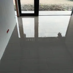 Błyszcząca, świeżo ułożona podłoga z płytek w jasnych odcieniach szarości, widoczne odbicie okna tarasowego z widokiem na ogród w tle, ściana z widocznymi puszkami elektrycznymi.