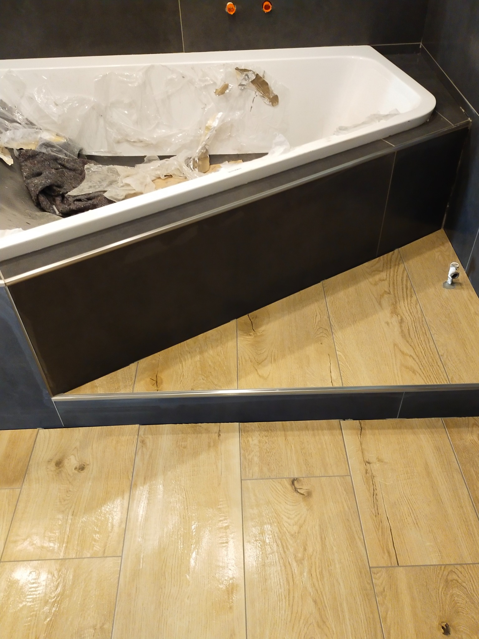 Wanna narożna zabezpieczona folią i kocem, obudowana ciemnymi płytkami, podłoga wyłożona jasnymi panelami imitującymi drewno, widoczne elementy instalacji wodnej.