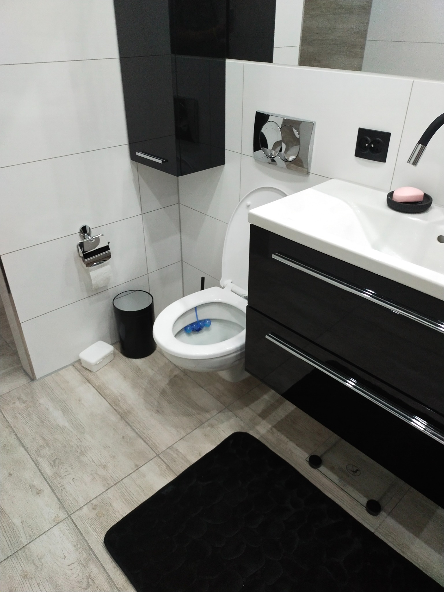 Nowoczesna łazienka z białymi płytkami, czarnymi szafkami i drewnopodobną podłogą, widoczny fragment toalety z niebieskim odświeżaczem i czarny dywanik.