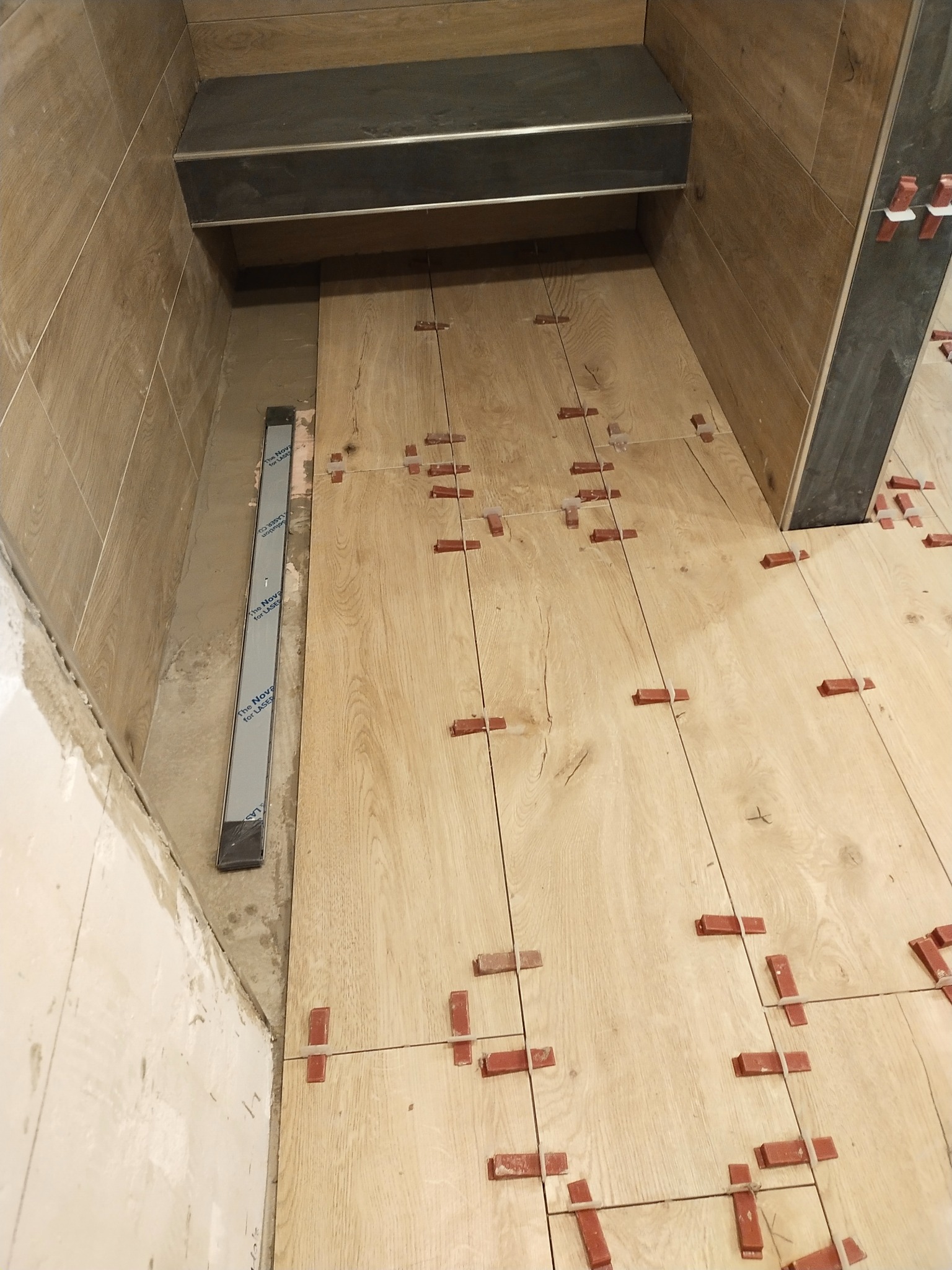 Układanie jasnych płytek imitujących drewno na podłodze w łazience z użyciem poziomicy laserowej i czerwonych krzyżyków dystansowych.