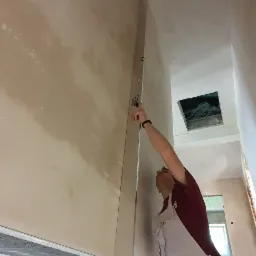 Pracownik w kombinezonie murarskim sprawdza pion ściany po nałożeniu tynku maszynowego za pomocą długiej poziomicy. Widoczne przewody elektryczne wystające ze ściany i otwór w suficie.