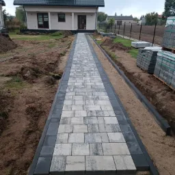 Ułożona ścieżka z szaro-białej kostki brukowej prowadząca do domu, z czarnym obramowaniem, widoczne palety z kostką i przygotowany teren pod trawnik.