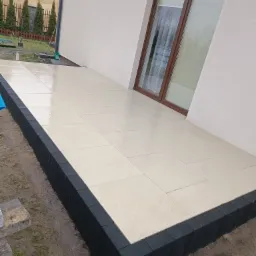 Nowo wyłożony taras z jasnych, kwadratowych płyt, obramowany ciemnoszarą kostką, przylegający do ściany domu z oknem.