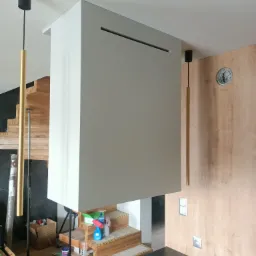 Biała zabudowa nad schodami z poziomym otworem wentylacyjnym, widoczne fragmenty drewnianych schodów, lampy wiszące w kształcie złotych rurek i drewniana boazeria ścienna.