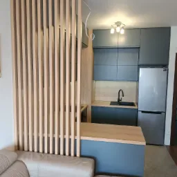 Nowoczesna kuchnia z szarymi szafkami, drewnianym blatem i ażurową ścianką działową z jasnego drewna, widoczny fragment beżowej sofy.