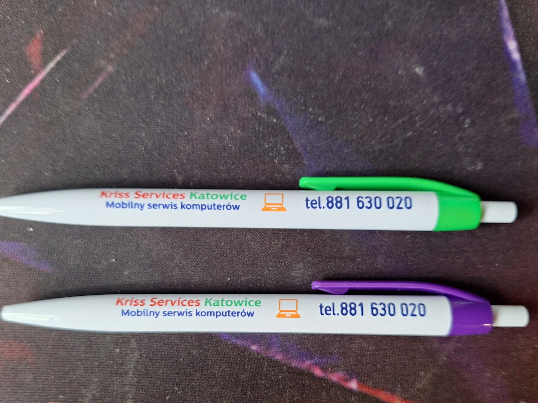 Dwa długopisy reklamowe firmy Kriss Services Katowice z nadrukowanym logo, adresem i numerem telefonu, leżące na ciemnym tle. Jeden długopis ma zielony klips, a drugi fioletowy.