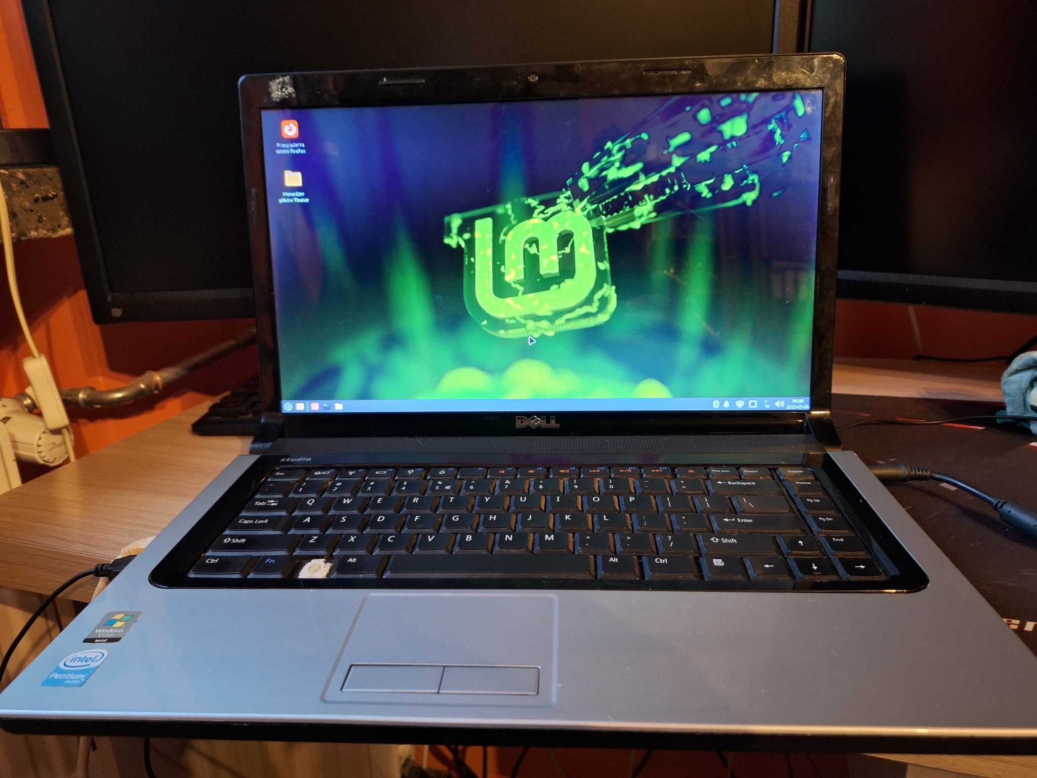 Laptop Dell Studio z systemem Linux Mint na ekranie, widoczne uszkodzenia klawiatury, zarysowania na obudowie. W tle monitory.