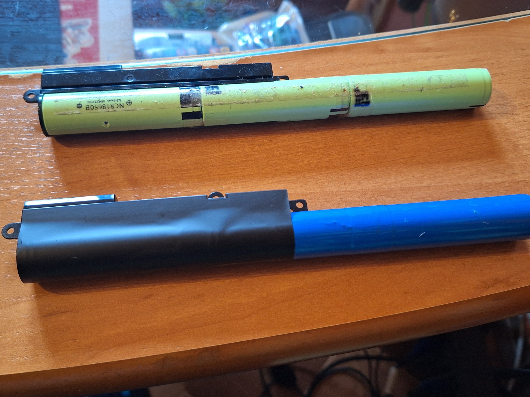 Dwie baterie litowo-jonowe, jedna zielona z oznaczeniem NCR18650B, druga niebieska z czarną obudową, leżące na drewnianym blacie. Zużyte ogniwa.