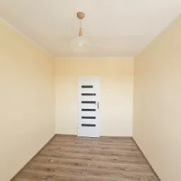 Pusty pokój po odświeżeniu: beżowe ściany, drewnopodobna podłoga, białe drzwi z czarnymi paskami i lampa z drewnianymi elementami.
