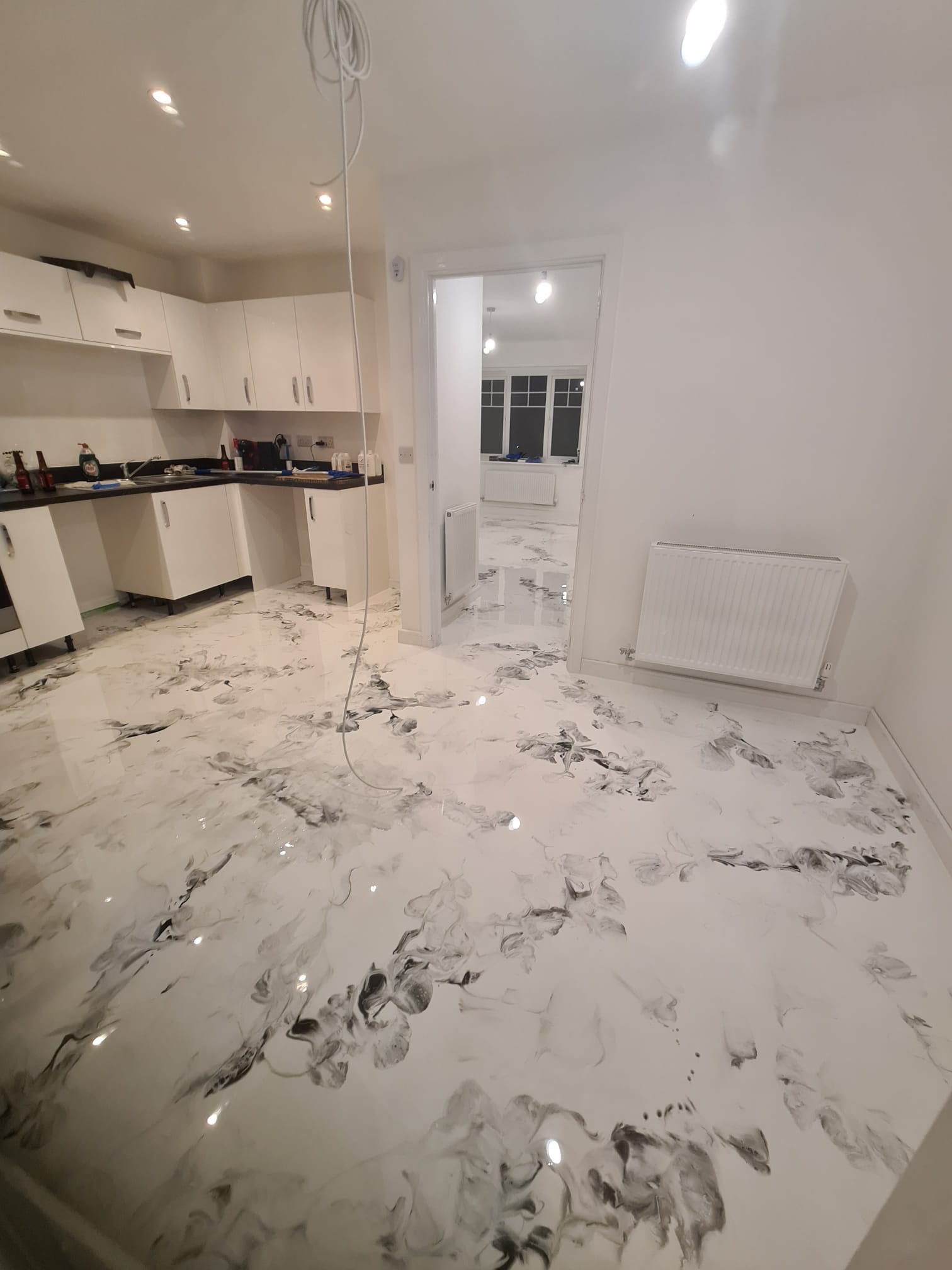 Wykończona podłoga żywiczna w odcieniach bieli i szarości z marmurkowym wzorem w kuchni i przyległym pomieszczeniu; białe szafki kuchenne i grzejnik na ścianie.