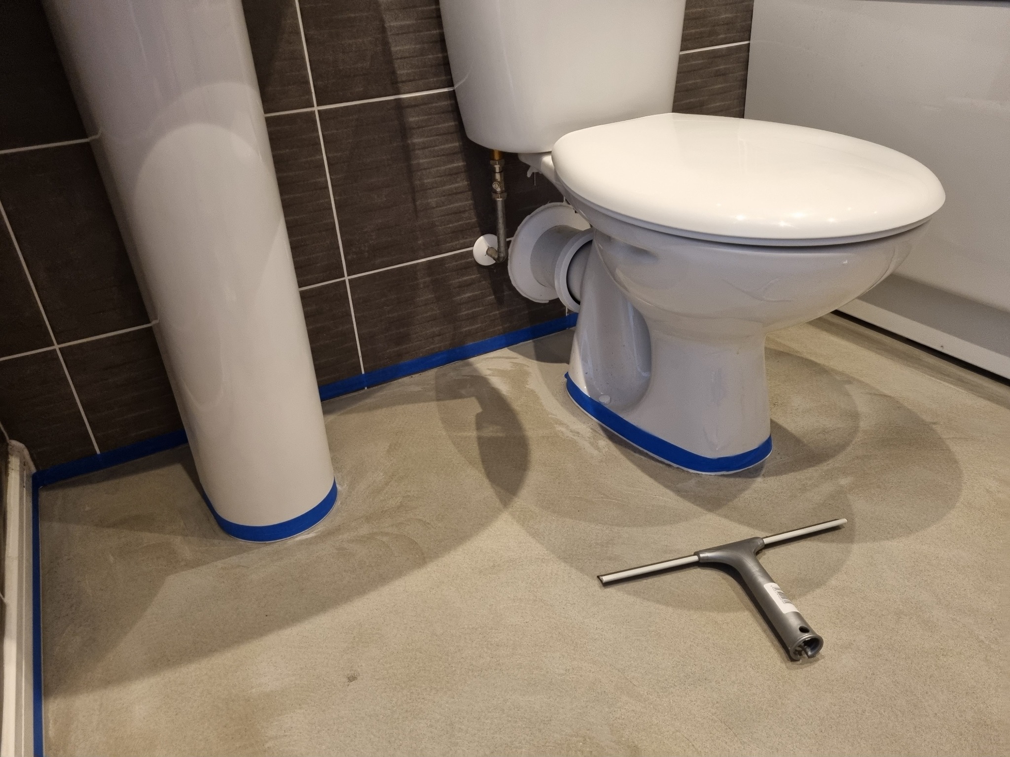 Przygotowanie do prac wykończeniowych w łazience w Rugby: zabezpieczona taśmą umywalka i toaleta na betonowej podłodze, obok leży ściągaczka do wody.