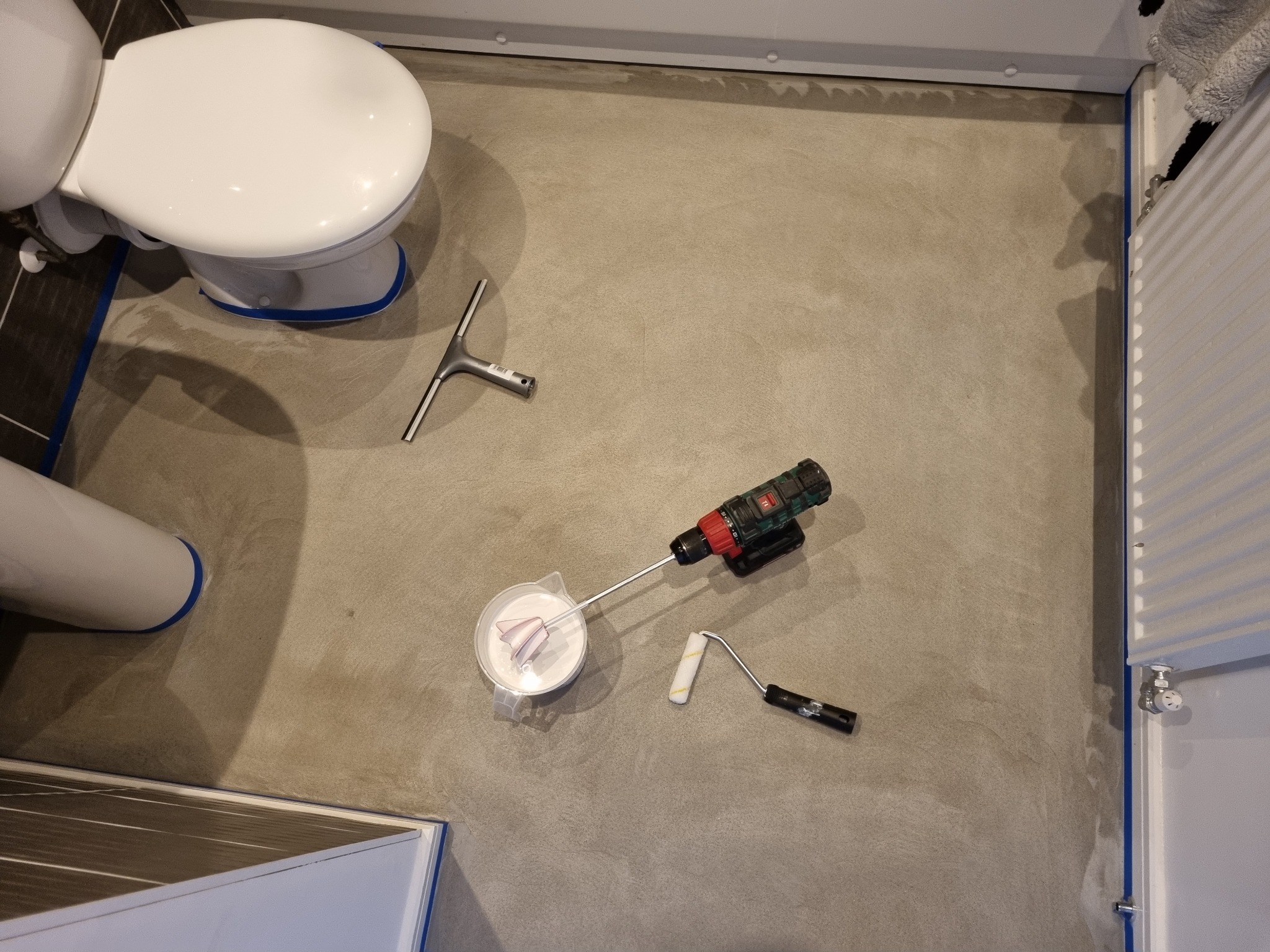 Przygotowanie do malowania podłogi w łazience: widoczny grunt w pojemniku z mieszadłem na wiertarce akumulatorowej, wałek do malowania oraz ściągaczka do wody, podłoga zabezpieczona taśmą malarską...