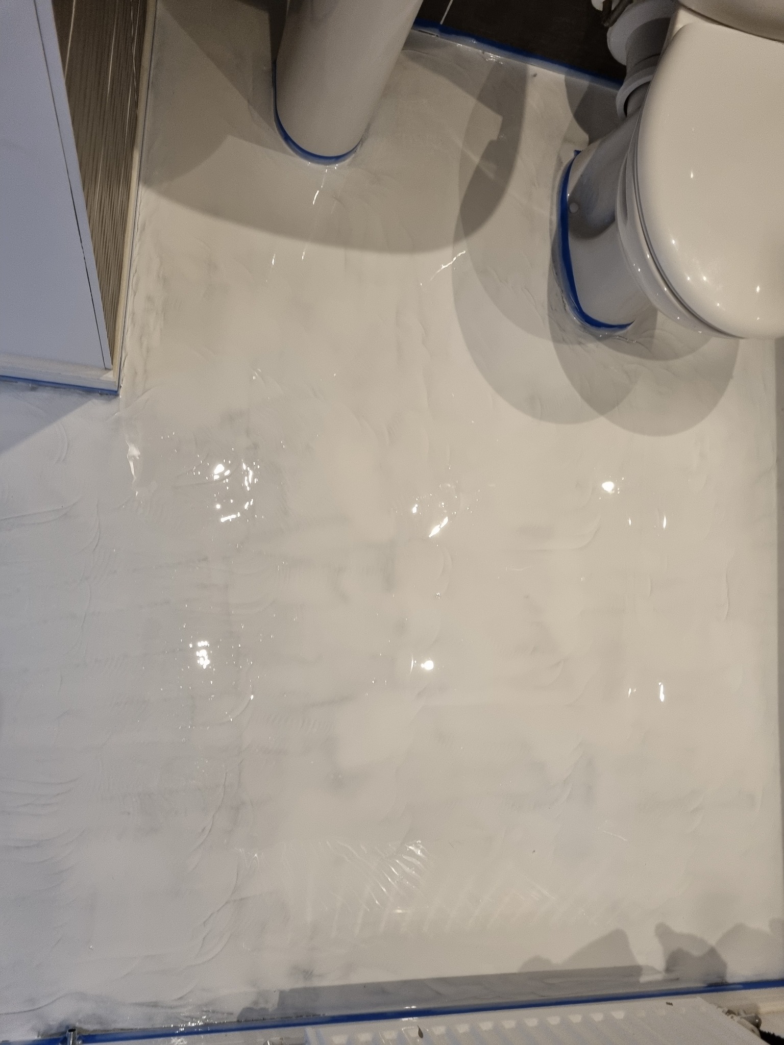 Biała, błyszcząca wylewka żywiczna na podłodze w łazience, zabezpieczona niebieską taśmą malarską przy krawędziach i wokół toalety, widoczne refleksy światła.