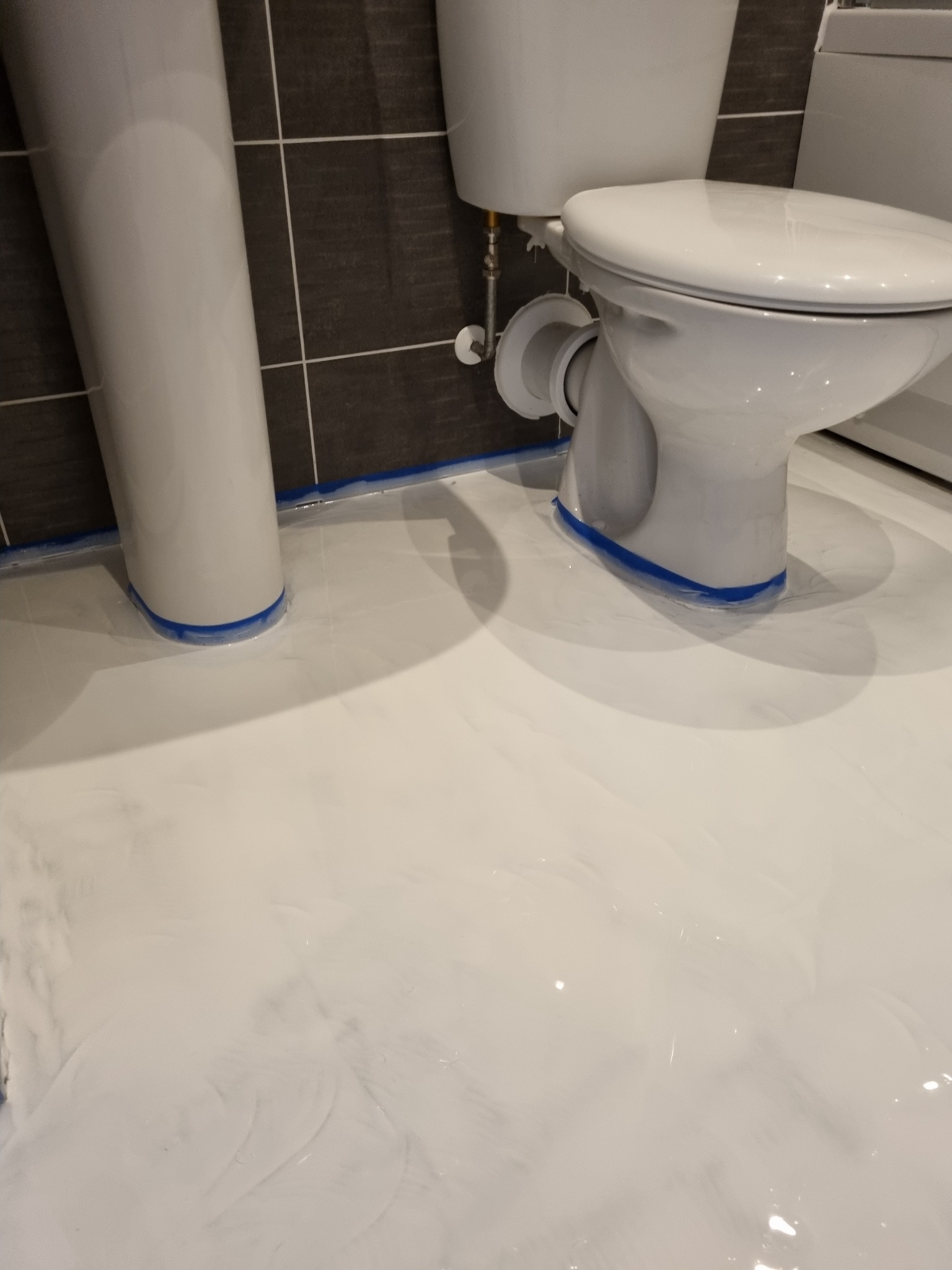 Biała, błyszcząca posadzka epoksydowa w łazience w trakcie wylewania, zabezpieczona niebieską taśmą malarską wokół toalety i umywalki.