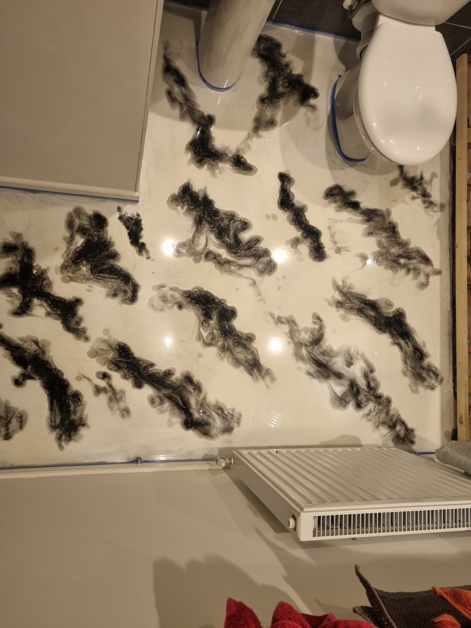 Biała posadzka żywiczna w łazience z abstrakcyjnym wzorem czarnych smug, widoczny fragment toalety, grzejnika i zabudowy, zabezpieczone niebieską taśmą malarską.