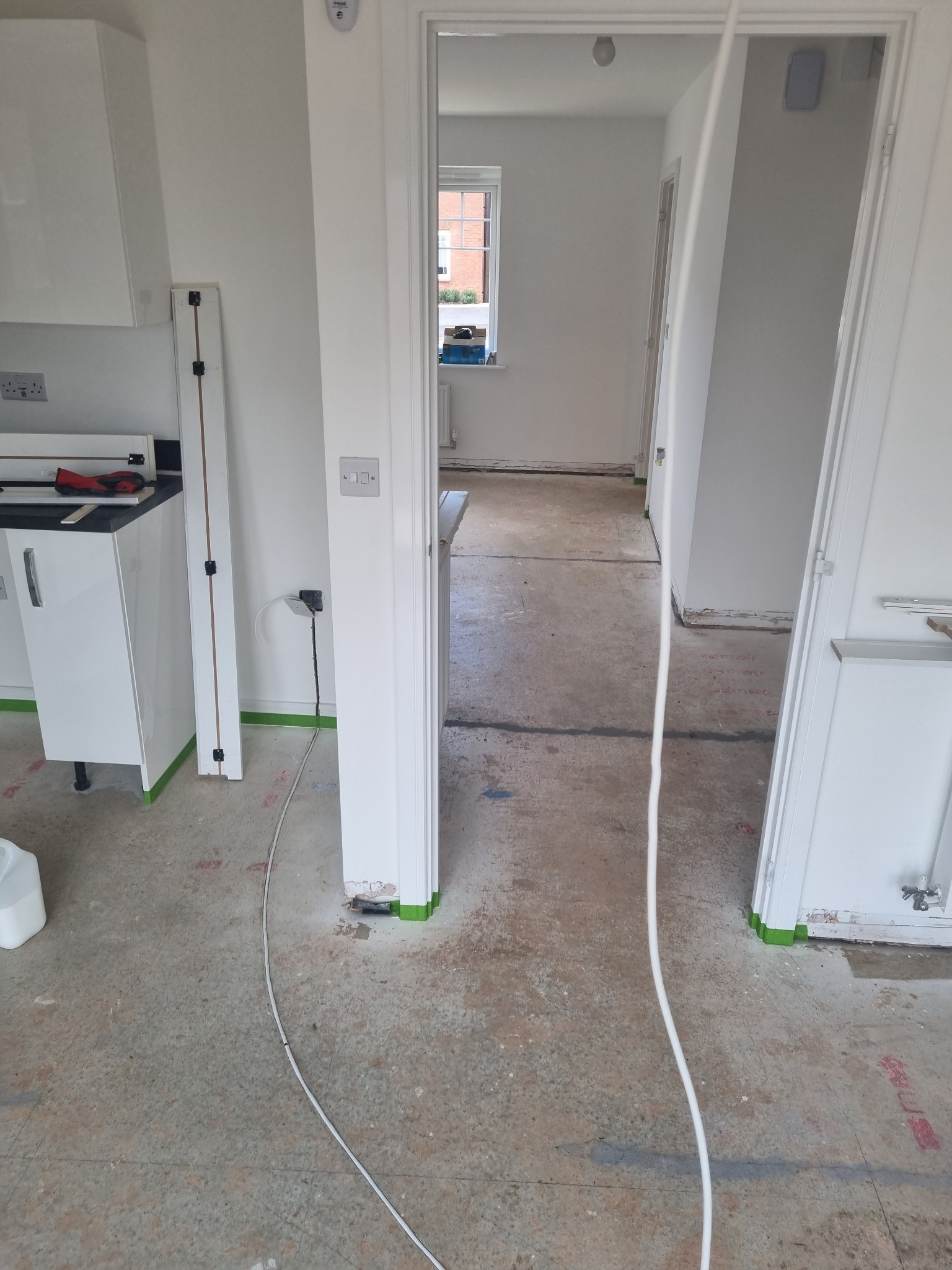 Wnętrze w trakcie prac wykończeniowych: białe ściany, częściowo zamontowane białe meble kuchenne, widoczne przewody elektryczne i taśma malarska zabezpieczająca podłogę, surowa podłoga przed...